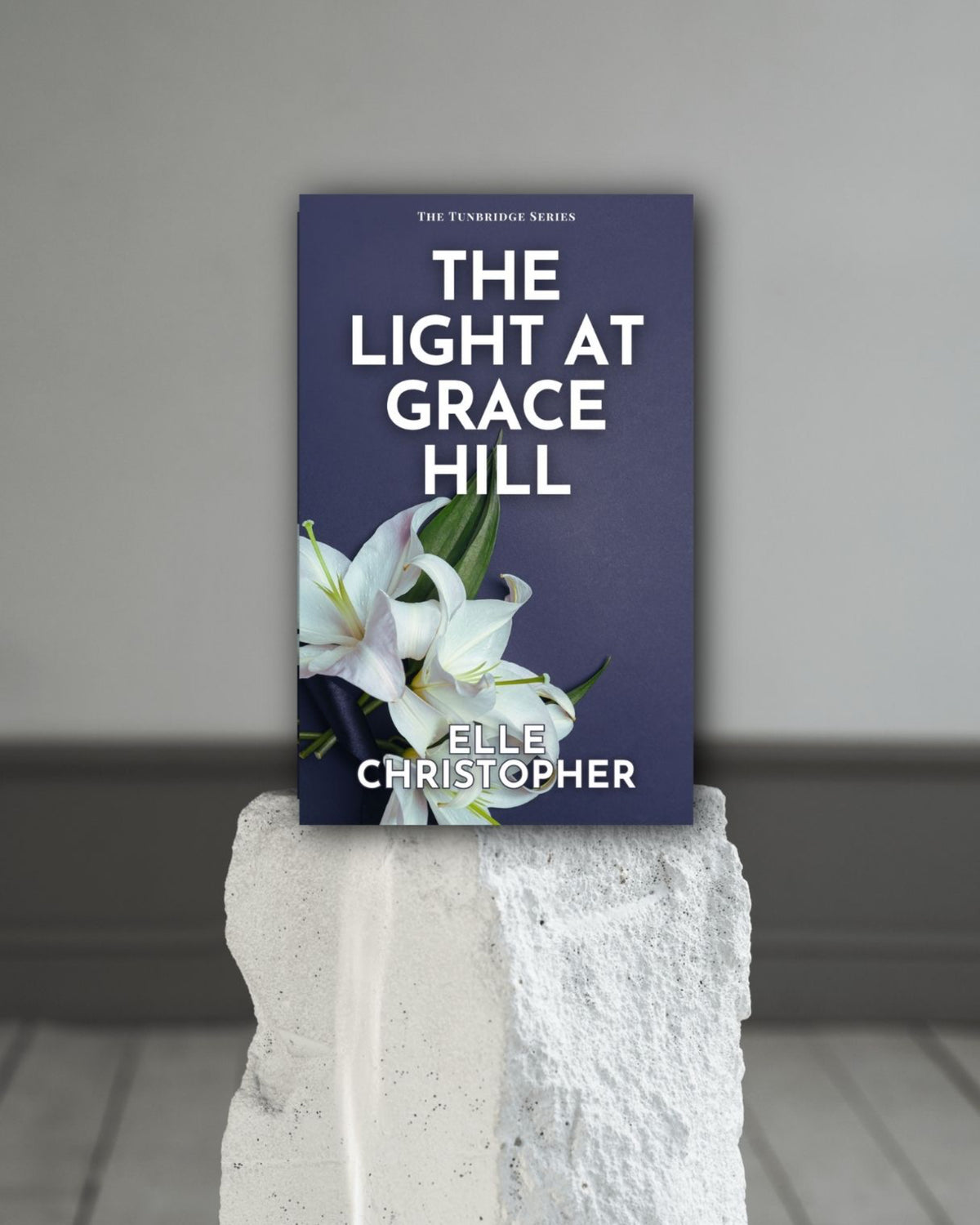 Book titled 'The Light at Grace Hill' by Elle Christopher on a white pedestal against a gray background